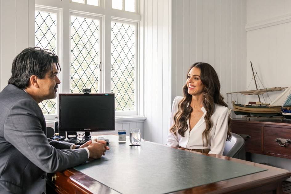 Two people are conversing at a desk in an office with a window and model ship in the background, discussing their options with family lawyers Brisbane.
