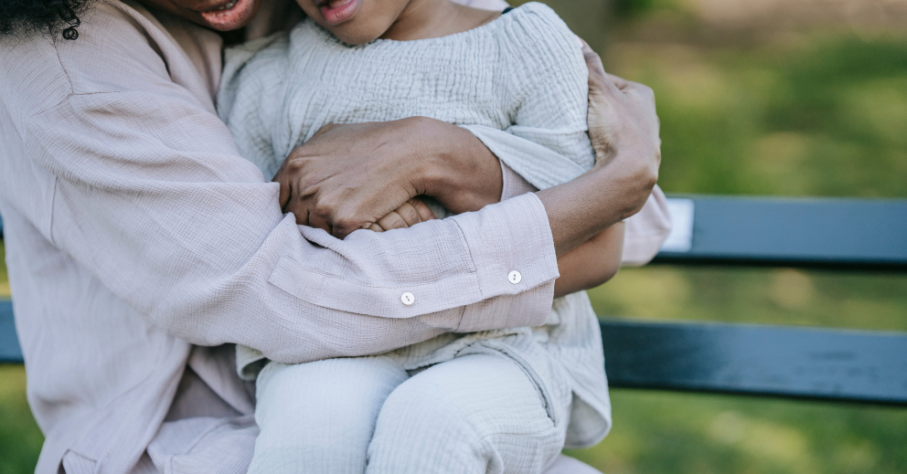 Adult and child in light clothes sitting together on a bench outdoors.