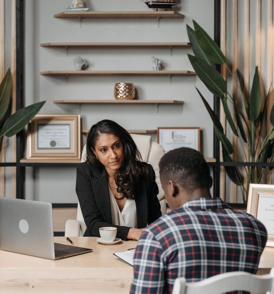 Two people having a conversation at a desk with a laptop and framed certificates.