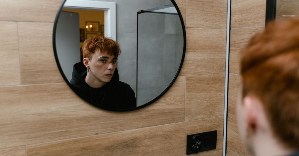 Person with short red hair looks at their reflection in a round bathroom mirror who seemed to have experienced domestic abuse
