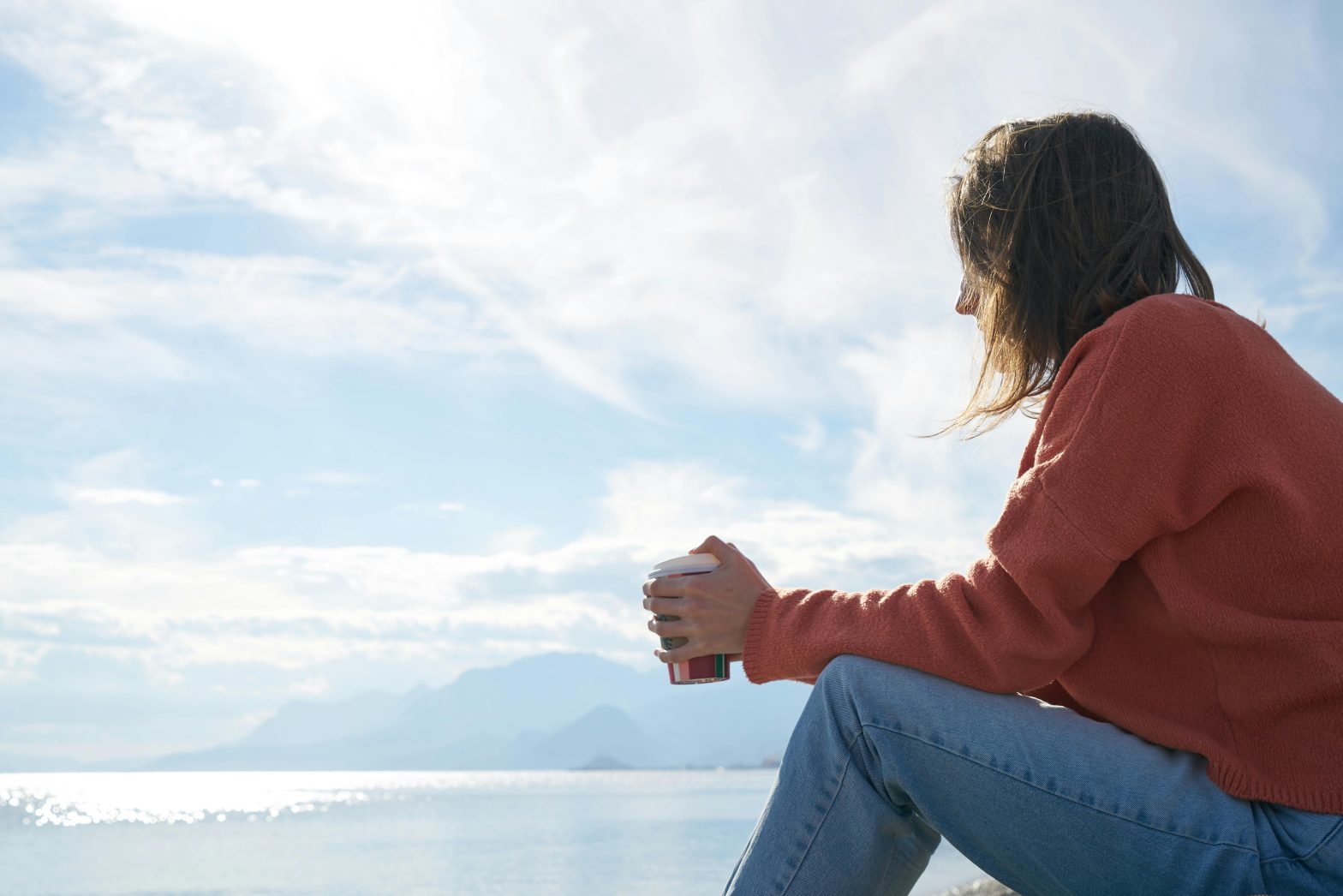 Person in a red sweater holds a cup while sitting by a calm sea under a cloudy sky.