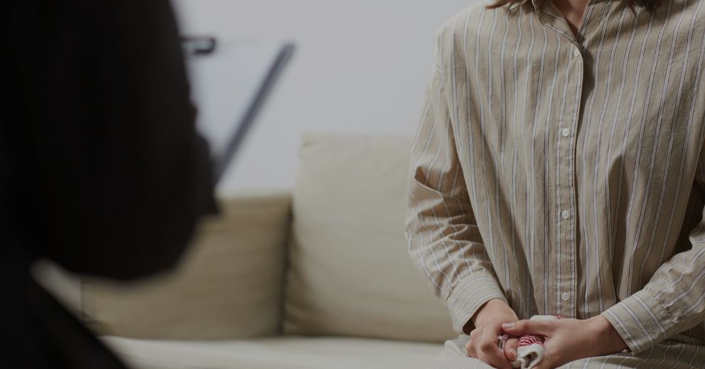 Person holding hands while sitting on a couch, facing someone with a clipboard out of focus.