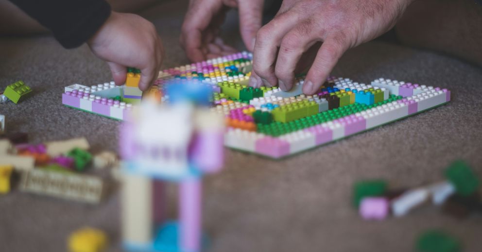 Hands assembling colorful plastic building blocks on a flat surface, with pieces scattered nearby.
