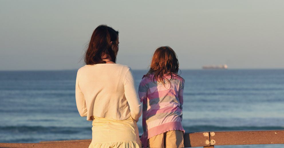 Two people, an adult and a child, stand by a wooden railing looking out at the ocean.