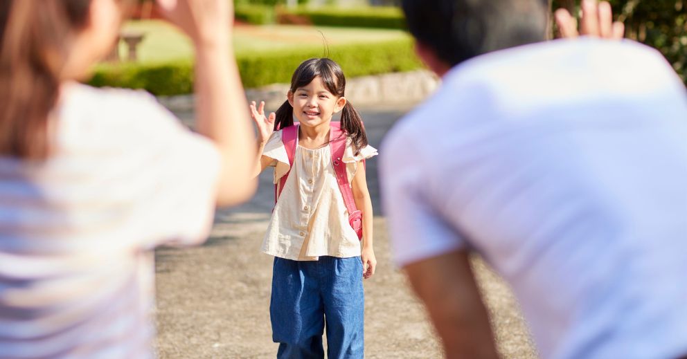 Young girl with a backpack waves while two adults stand in the foreground, waving back.