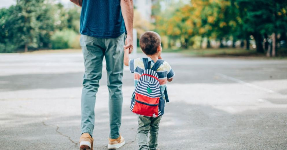 Adult and child with backpack walking outdoors on a paved path, seen from behind.