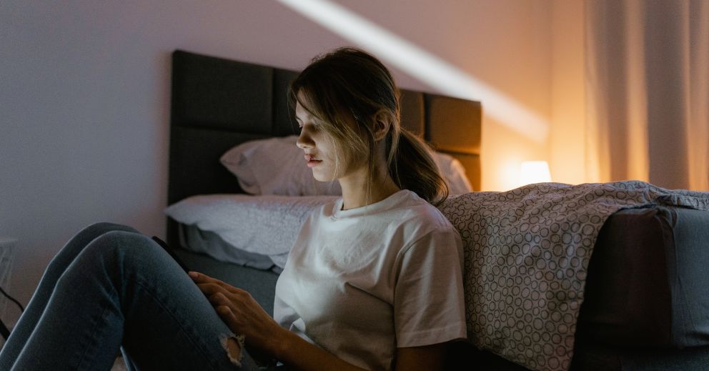 Woman sitting on floor by bed, using a tablet or phone in a dimly lit bedroom.