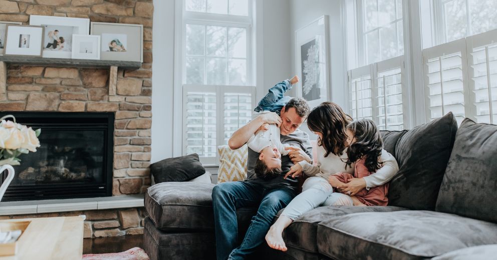 Family of four laughing and playing together on a gray sectional sofa in a living room.