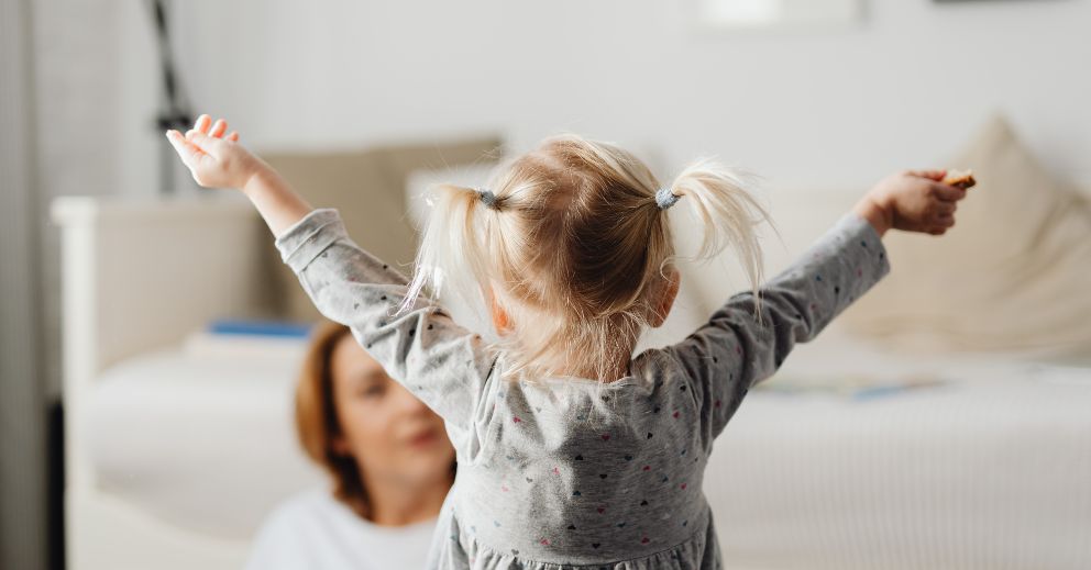 Young child with pigtails raises arms while an adult sits in the background at home.