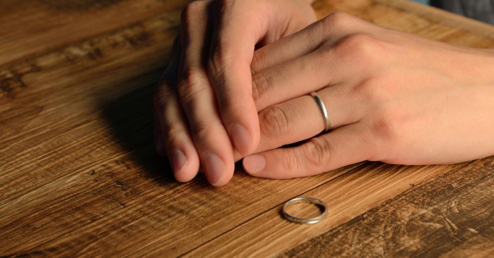Hands on a wooden table with a wedding ring removed and placed beside them.