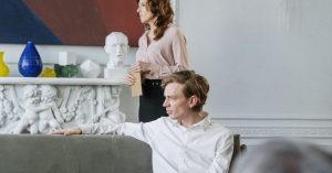 Man sitting on a couch while a woman stands behind him holding an envelope for spousal maintenance.