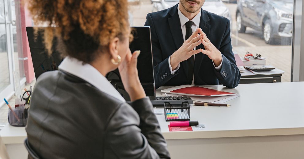 Two people in business attire sit across from each other at a desk in an office setting.
