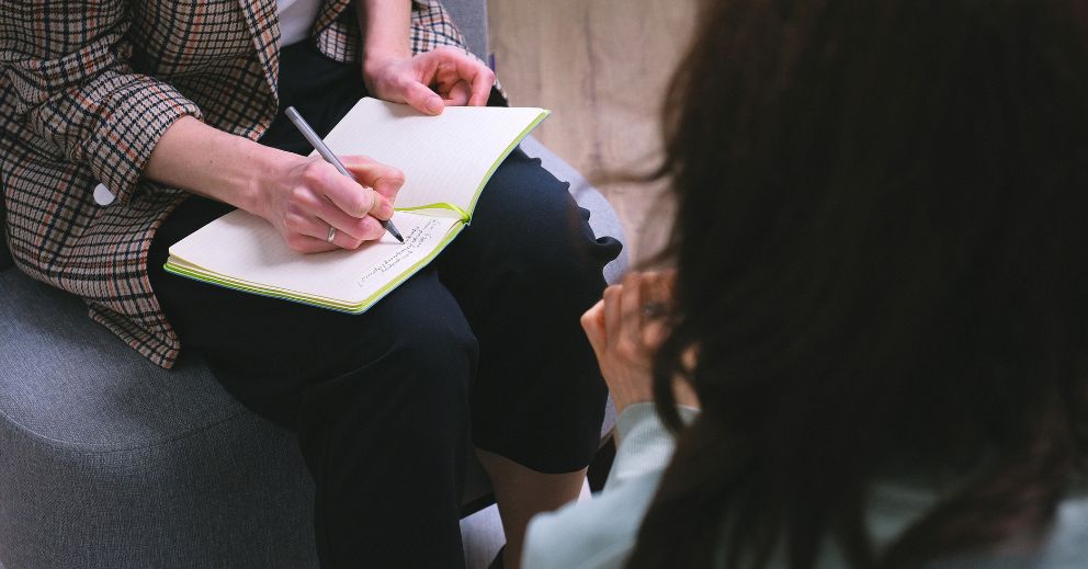 Two people sit; one writes notes in a notebook while the other listens or speaks.